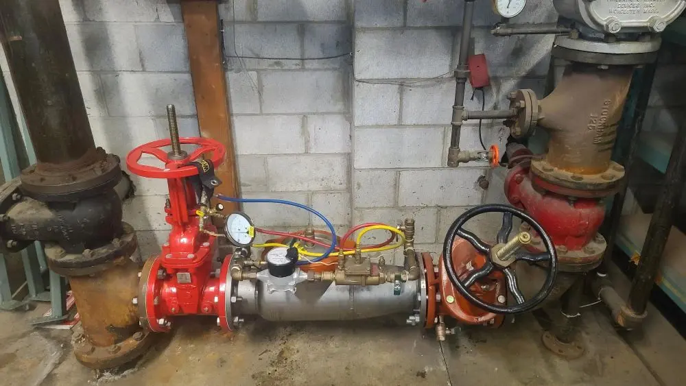 A backflow prevention assembly with red and black valves, a gauge, and blue, red, and yellow hoses is shown against a cinder block wall.