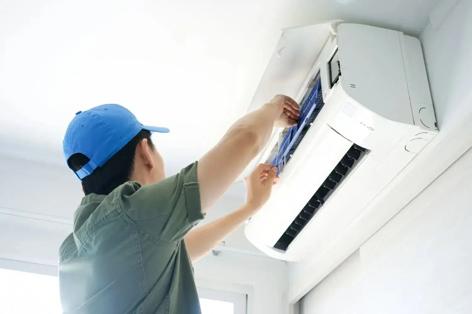 A male technician is wearing a blue cap and a gray shirt and is working on the internal components of a white wall-mounted air conditioning unit.