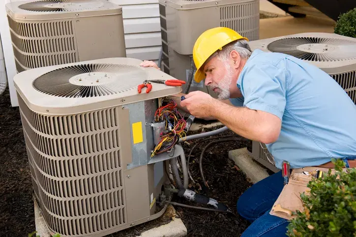 An HVAC technician in a yellow hard hat is working on an outdoor air conditioning unit.