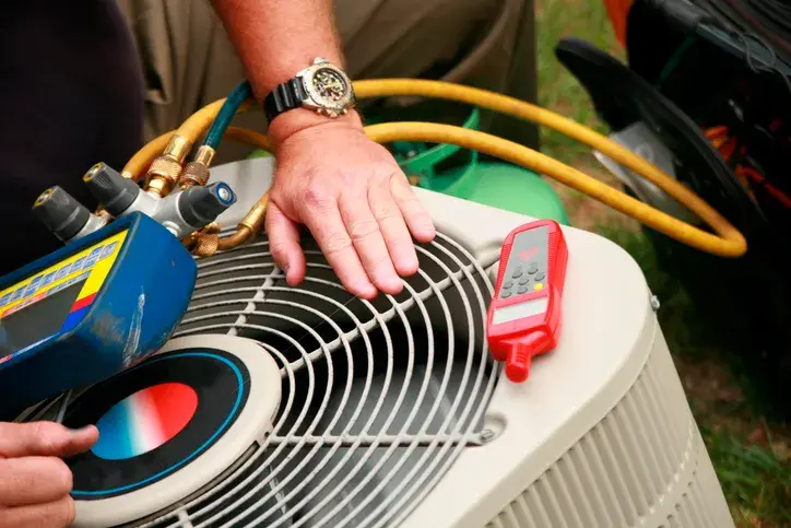 A person is using HVAC diagnostic tools, including a manifold gauge and a red remote, on an outdoor air conditioning unit.