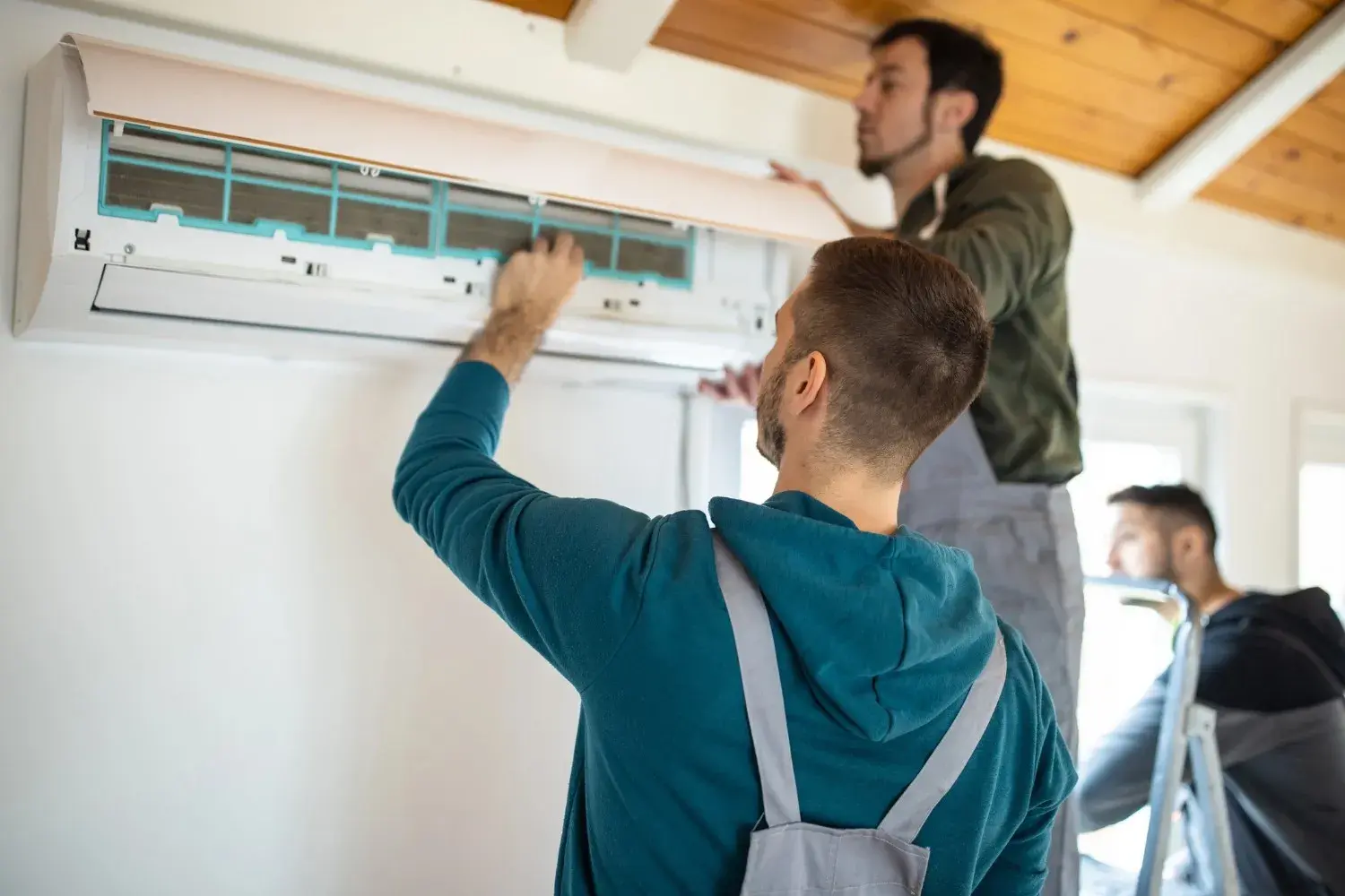 Three technicians are installing a wall-mounted ductless air conditioning unit in a room with a sloped wooden ceiling.