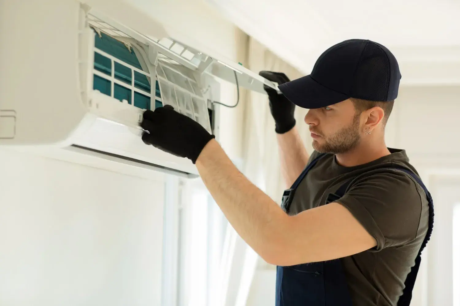 A male technician in a black cap and gloves is servicing a wall-mounted ductless air conditioning unit.