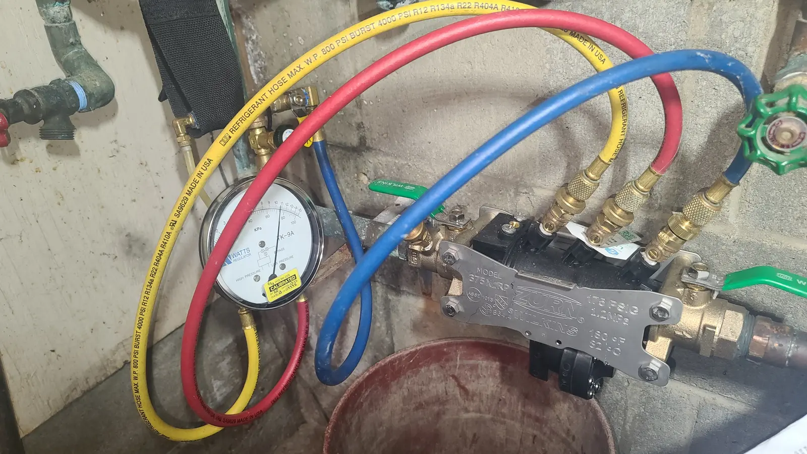 A plumber is performing a backflow test with a gauge and a system of red, yellow, and blue hoses attached to copper and brass pipes.