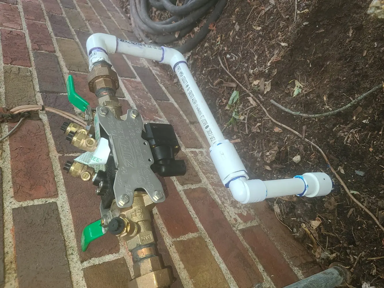 A Zurn backflow preventer is connected to white PVC pipes and brass valves, installed on the exterior of a brick wall.