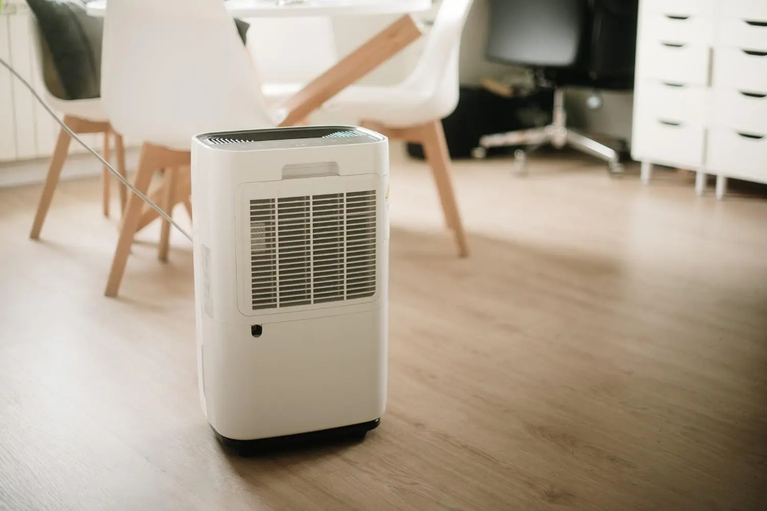  A single white portable dehumidifier stands on a light wood floor in a clean, modern room. The dehumidifier has a large black grille on the side and a handle on top. In the background, out of focus, are a white dining table with white chairs and a white chest of drawers.
