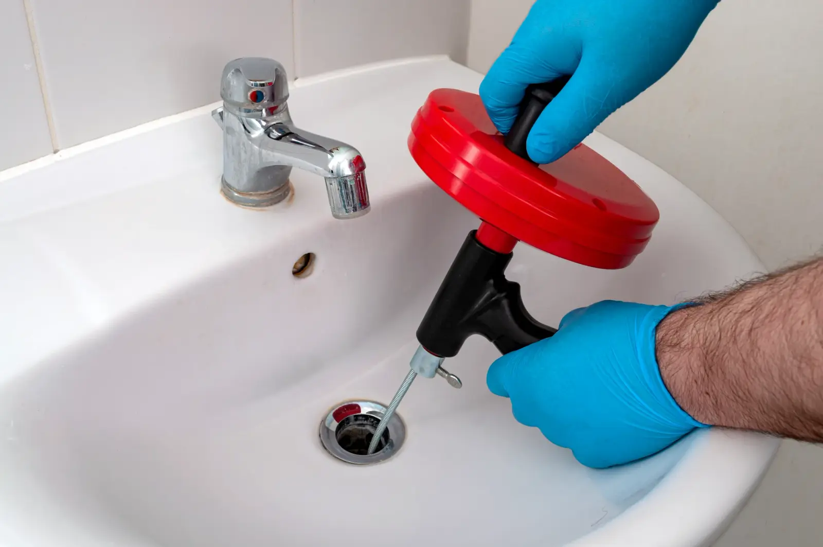 Person in blue gloves uses a red drain snake to clear a white bathroom sink's clogged drain.