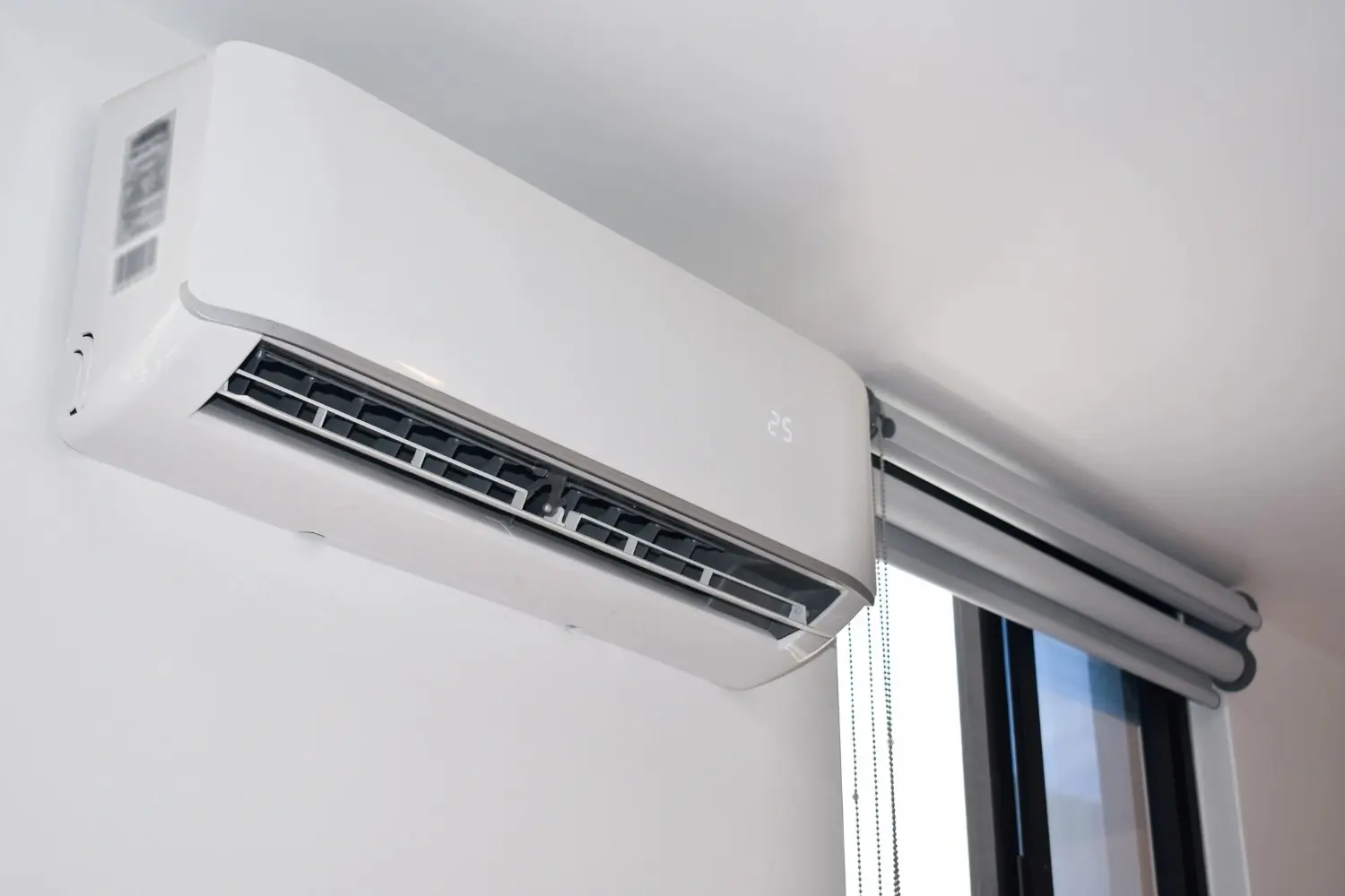 A sleek, white ductless mini-split air conditioner is mounted on a white wall near a window with a roller shade.