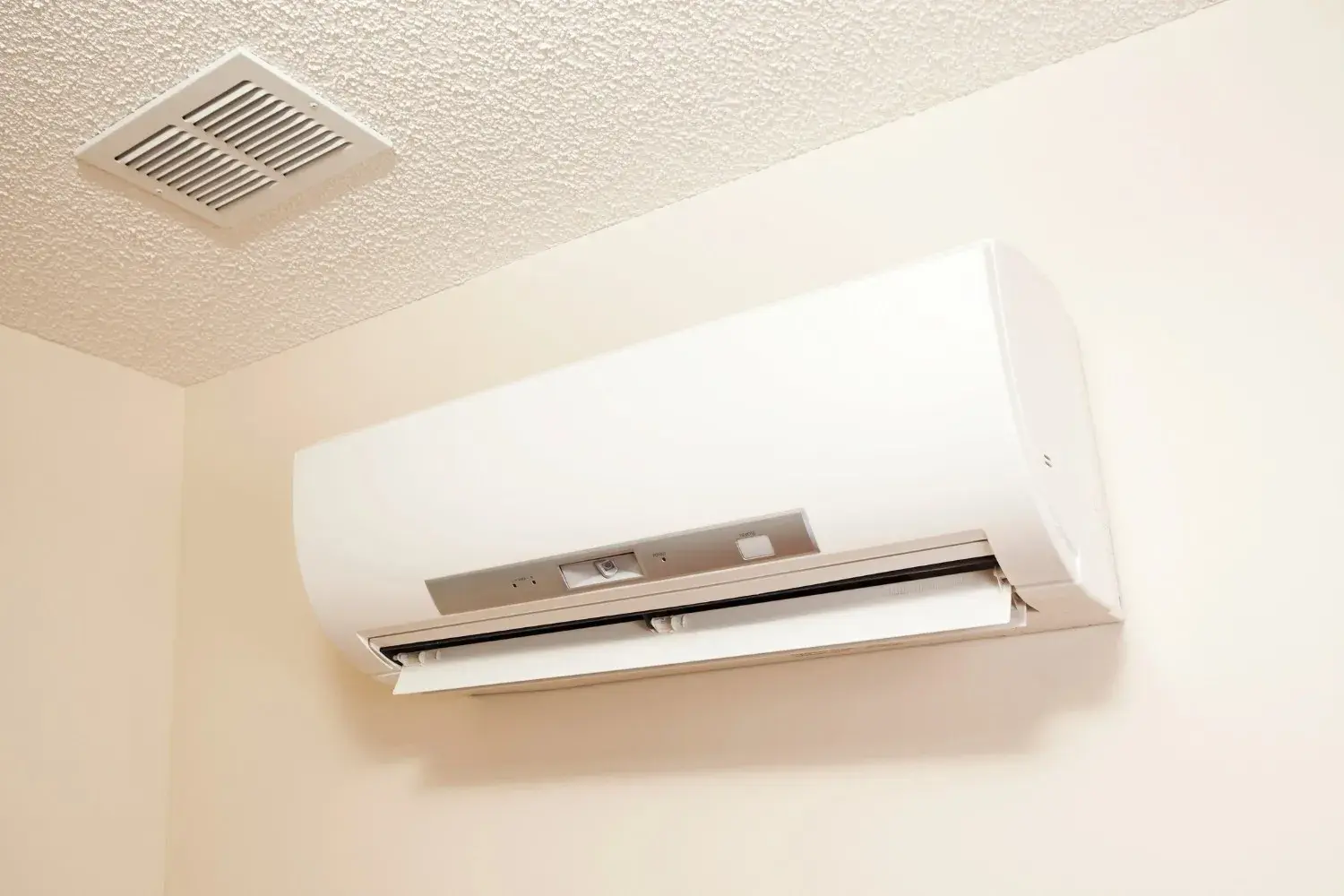 A white, ductless mini-split air conditioning unit is mounted on a light-colored wall with a ceiling vent above it.