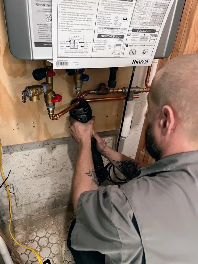 A plumber with a beard and tattoos is working with a power tool on the copper pipes of a wall-mounted Rinnai water heater.