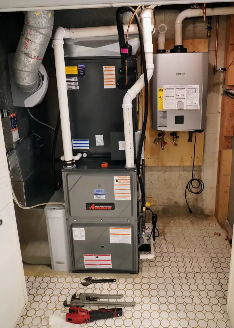 A vertical gray furnace is installed in a mechanical room next to a Rinnai tankless water heater, with tools on the tiled floor.
