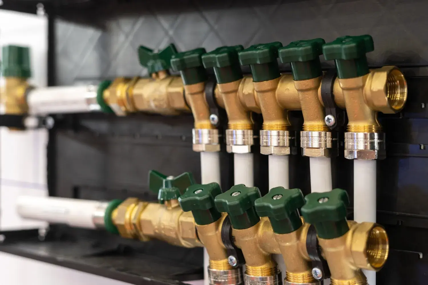 A close-up of a plumbing manifold shows a series of brass valves with green handles connected to white pipes.