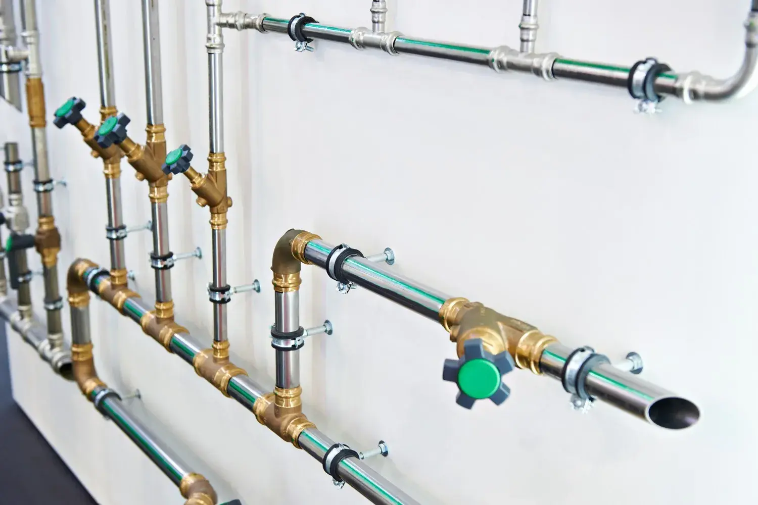 A clean and complex network of metal pipes, valves, and fittings is mounted on a white wall.