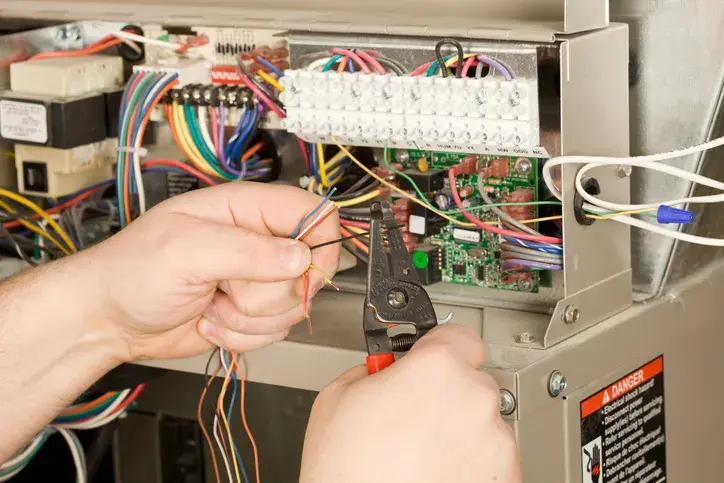 A technician's hands are shown using wire strippers to work on the complex wiring of a furnace control board.