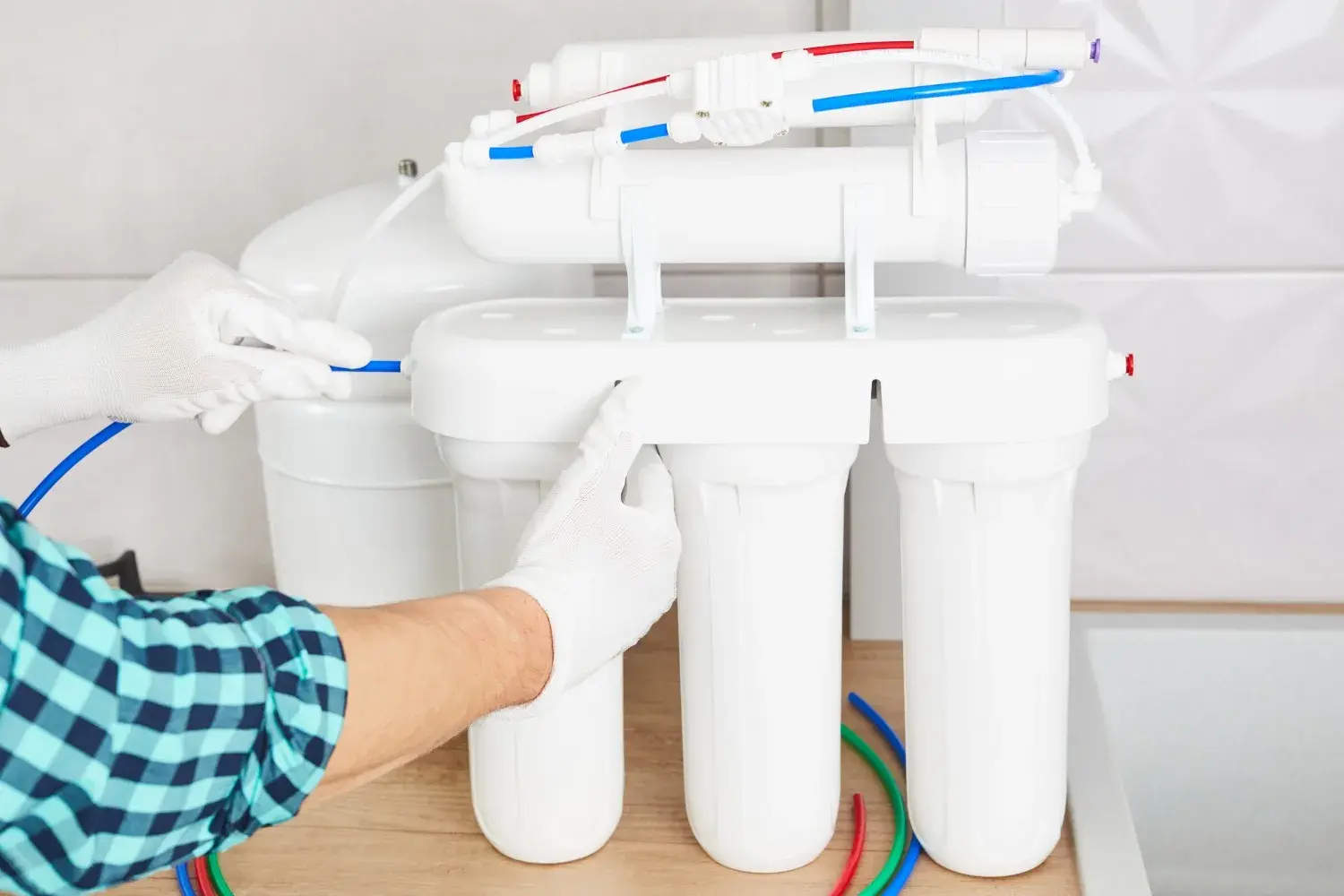 A gloved plumber in a checkered shirt is installing a reverse osmosis water filtration system with multiple white filters and red and blue plastic tubing.