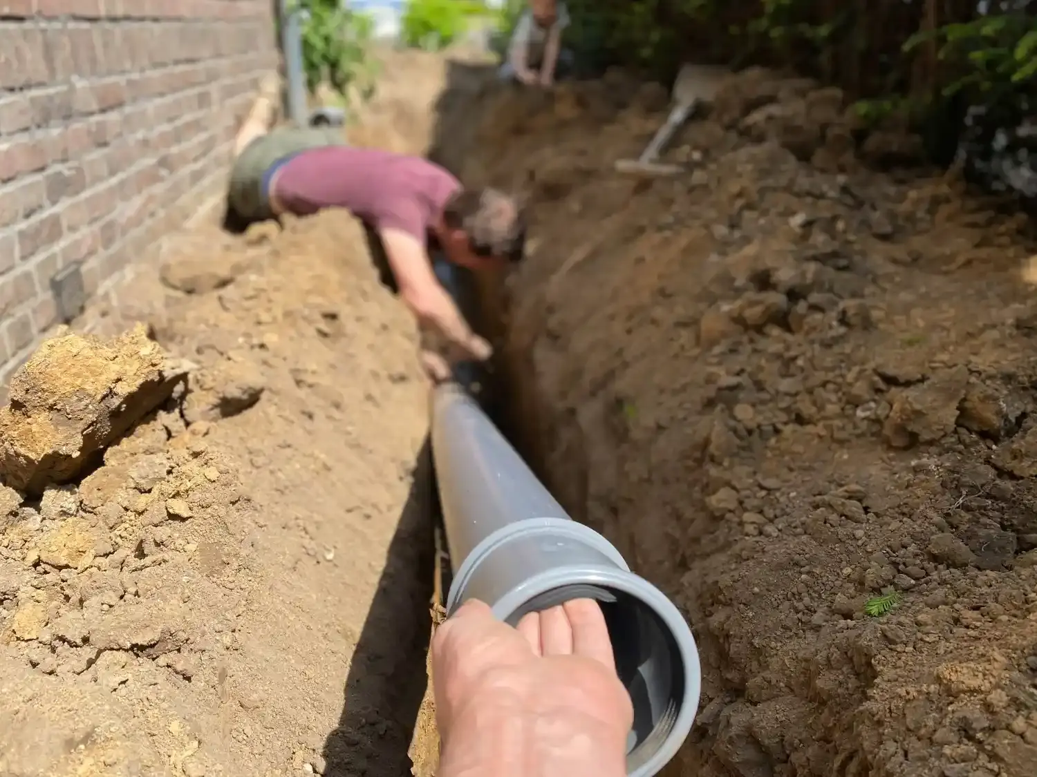 A plumber is installing a large gray sewer pipe in a long trench dug in the ground next to a brick house.
