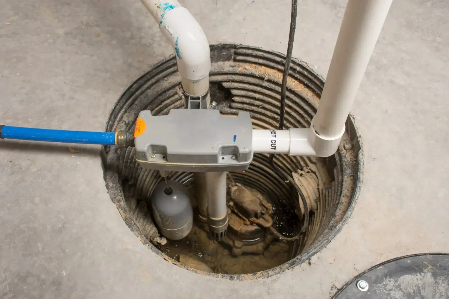 A top-down view of a sump pump installed in a basement pit, with various white and blue pipes leading to a gray control box and a floating gray sensor.