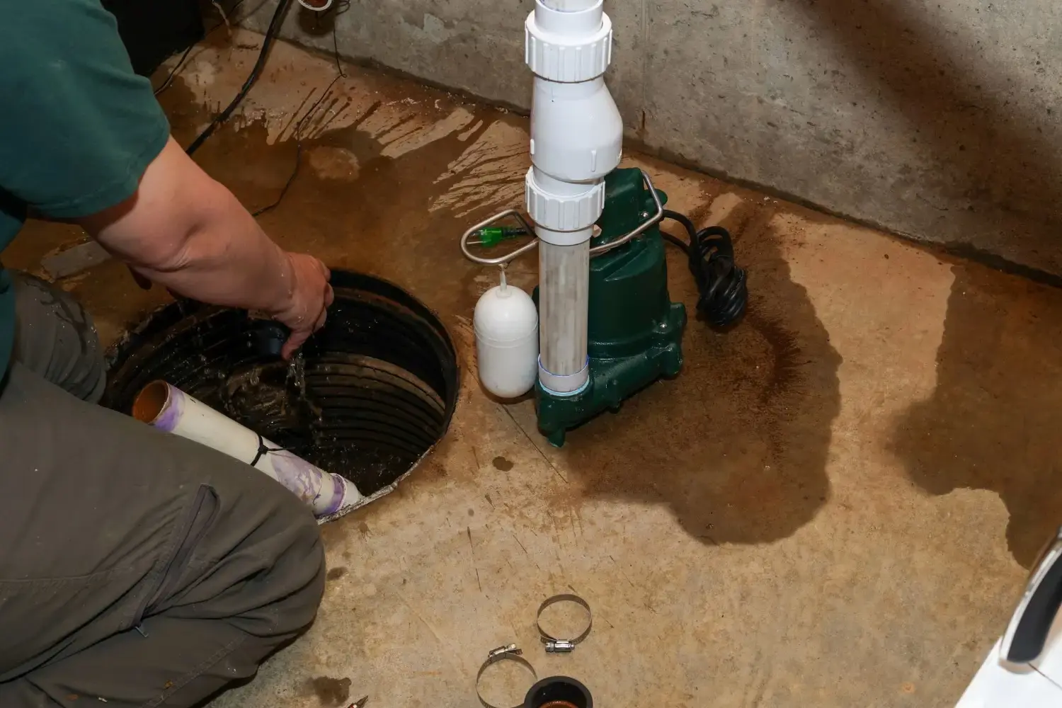 A male plumber is working on a sump pump, with its white and blue PVC pipes being installed in a basement.