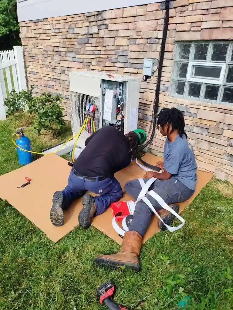 Two HVAC technicians are kneeling on cardboard sheets on a lawn, working on an outdoor mini-split air conditioning unit.
