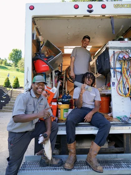 Three Sureway technicians posing at the back of a work truck loaded with tools, hoses, and equipment. One kneels giving a thumbs-up, one sits on the truck bed, and one stands in the cargo area.